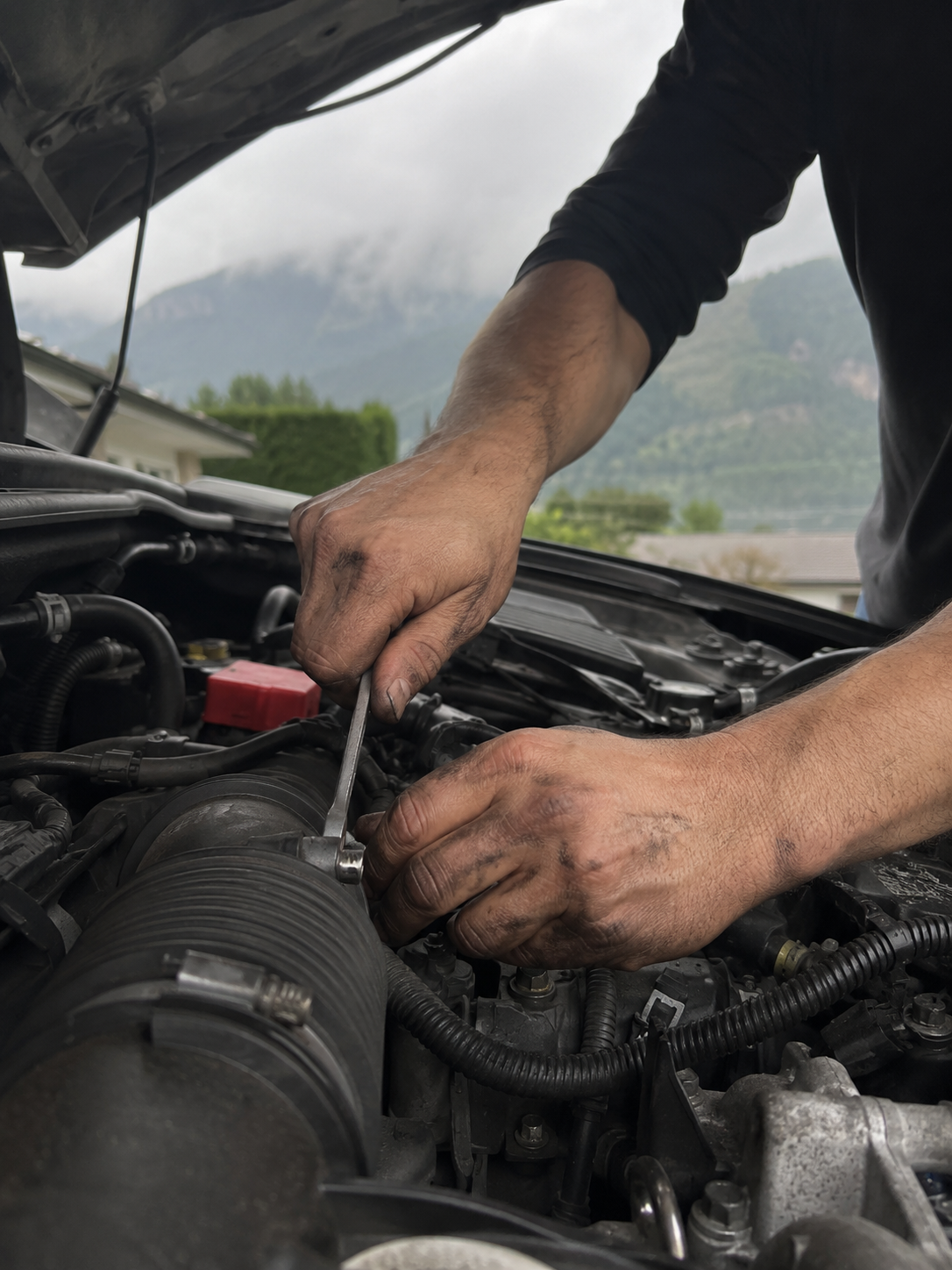 Close-up of a mobile mechanic's hands using a wrench under the hood of a car with North Vancouver mountains in the background